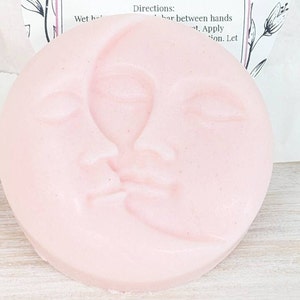 May include: A round, pale pink soap bar featuring a molded relief design of a crescent moon and a human face in profile. The surface is smooth with subtle texture. Behind the soap, a partial white paper label displays the text Directions: Wet hair, rub bar between hands, apply to hair, let.