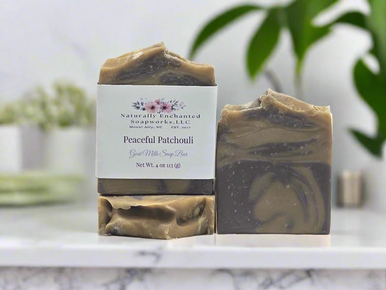 May include: Two bars of Peaceful Patchouli goat milk soap with a marbled brown and dark gray swirl pattern. One bar is wrapped in a white paper label featuring floral graphics and the text Naturally Enchanted Soapworks, LLC, Net Wt. 4 oz.
