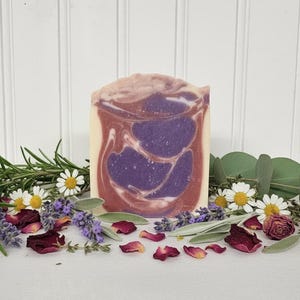 May include: A rectangular bar of soap featuring a marbled swirl pattern in shades of deep purple, dusty rose, and cream. The soap is surrounded by sprigs of fresh green rosemary, lavender buds, white chamomile flowers, and dried red rose petals.