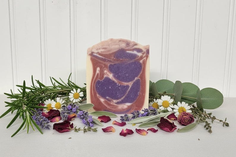 May include: A rectangular bar of soap featuring a marbled swirl pattern in shades of deep purple, dusty rose, and cream. The soap is surrounded by sprigs of fresh green rosemary, lavender buds, white chamomile flowers, and dried red rose petals.