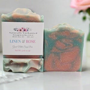 May include: Two bars of goat milk soap with a marbled pattern of teal, pink, and white. One bar is wrapped in a white paper label reading Naturally Enchanted Soapworks, L.L.C., Mount Airy, NC, EST. 2017, LINEN & ROSE, Goat Milk Soap Bar, Net Wt. 4 oz 113 (g).