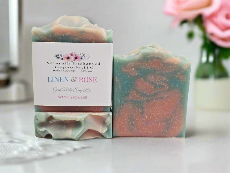 May include: Two bars of goat milk soap with a marbled pattern of teal, pink, and white. One bar is wrapped in a white paper label reading Naturally Enchanted Soapworks, L.L.C., Mount Airy, NC, EST. 2017, LINEN & ROSE, Goat Milk Soap Bar, Net Wt. 4 oz 113 (g).