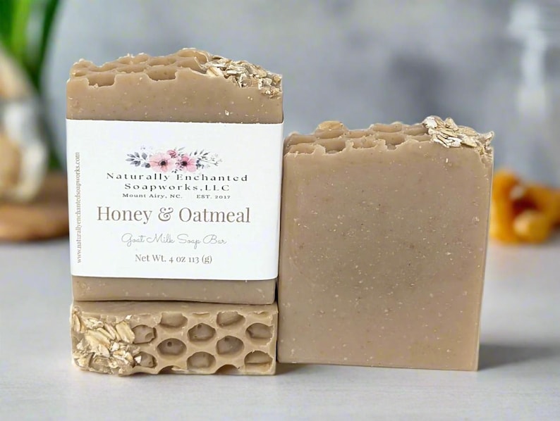 May include: A light brown Honey & Oatmeal goat milk soap bar with a honeycomb texture on top and embedded oats. The label reads Naturally Enchanted Soapworks, LLC, Honey & Oatmeal, Goat Milk Soap Bar, Net Wt. 4 oz 113 (g).