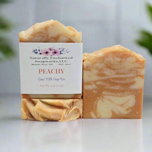 May include: Two rectangular bars of peach and cream swirled goat milk soap. One bar features a white paper label with floral graphics and text reading Naturally Enchanted Soapworks, LLC, PEACHY, Goat Milk Soap Bar, Net Wt. 4 oz (113 g).