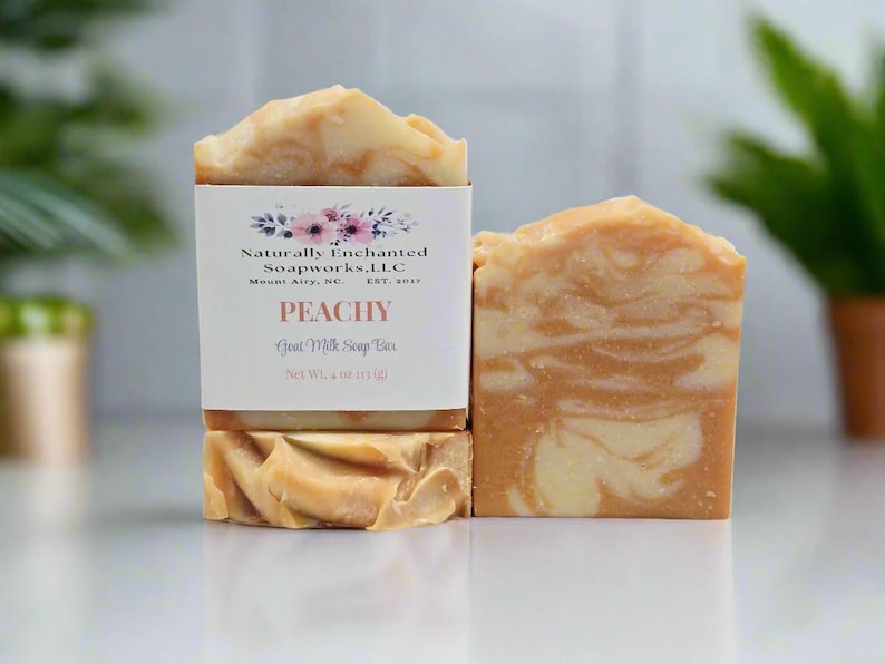 May include: Two rectangular bars of peach and cream swirled goat milk soap. One bar features a white paper label with floral graphics and text reading Naturally Enchanted Soapworks, LLC, PEACHY, Goat Milk Soap Bar, Net Wt. 4 oz (113 g).