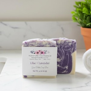 May include: A rectangular bar of soap with a purple and white marbled pattern. A white paper label wraps around the center, featuring floral graphics and the text: Naturally Enchanted Soapworks, L.L.C., Lilac + Lavender, Goat Milk Soap Bar, Net Wt. 4 oz.