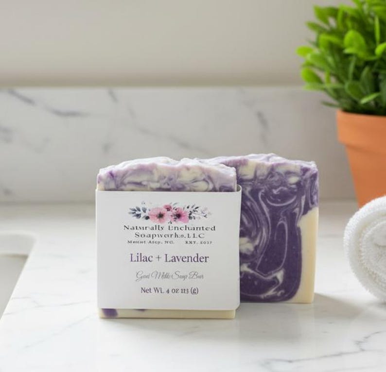May include: A rectangular bar of soap with a purple and white marbled pattern. A white paper label wraps around the center, featuring floral graphics and the text: Naturally Enchanted Soapworks, L.L.C., Lilac + Lavender, Goat Milk Soap Bar, Net Wt. 4 oz.