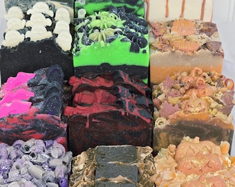 May include: A collection of twelve rectangular artisan soap bars arranged in a grid. The soaps feature various seasonal designs including ghosts, pumpkins, bats, and tombstones in colors of black, green, orange, purple, and white with glitter.