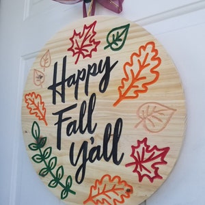 K&ouml;nnte beinhalten: Runder Holzschild mit dem Schriftzug "Happy Fall y'all" in schwarzer Schrift. Verziert mit bunten Bl&auml;ttern in Rot, Orange, Gr&uuml;n und Beige. Ein gr&uuml;nes Band und eine bordeauxfarbene Schleife sind zum Aufh&auml;ngen befestigt.