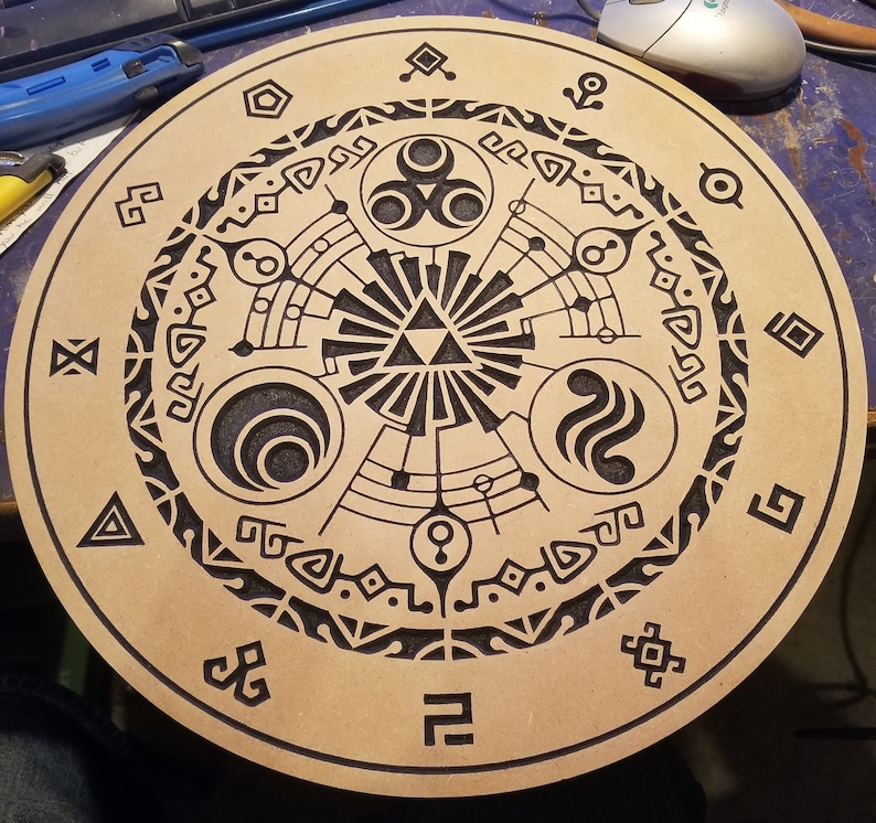 May include: A round, light brown wooden item with a detailed black engraved design. The design features geometric shapes, symbols, and a central triangle. The item appears to be a decorative piece or a game-related item.
