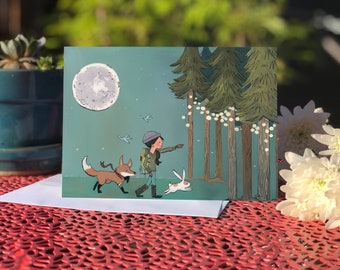 May include: A rectangular greeting card featuring a colorful illustration of a person with a backpack, a fox, and a rabbit walking through a forest at night under a full moon. The trees are decorated with glowing string lights and small birds fly nearby.