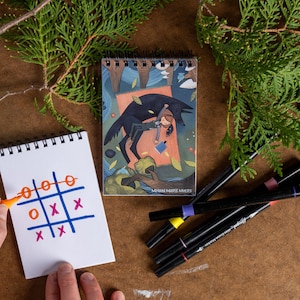 May include: A small, spiral bound notebook with a colorful illustration of a girl riding a wolf through a forest. The notebook has the text "Meghan Marie Myers" printed on the bottom. The notebook is lying on a brown surface with a few colorful markers next to it.
