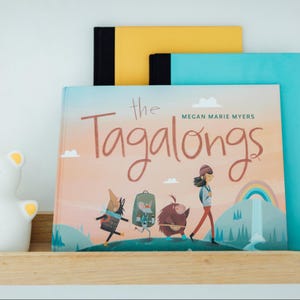May include: A children's book titled "The Tagalongs" by Megan Marie Myers. The cover features illustrations of cartoon characters, a rainbow, and a waterfall. The book is on a wooden shelf with other books.