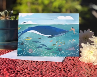 May include: A vibrant art print depicting a blue whale swimming in the ocean, surrounded by various marine life. A person is reaching out to touch the whale. The artwork features a light blue sky with clouds and birds.