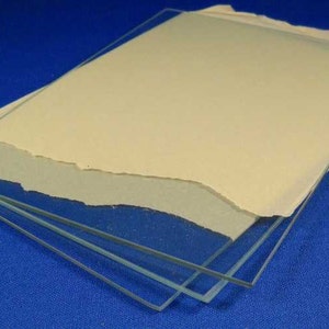 May include: Three clear glass sheets stacked on top of each other. The top sheet has a piece of beige paper underneath it.