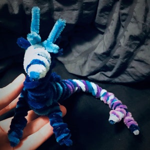May include: A blue and purple pipe cleaner dragon with a white striped head and three horns. The dragon is curled up in a hand.