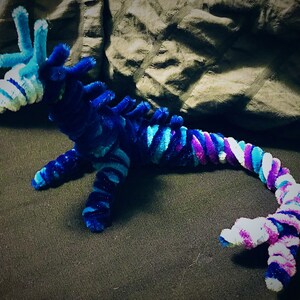May include: A blue, purple, and white pipe cleaner dragon with a long tail. The dragon is curled up on a dark gray surface.