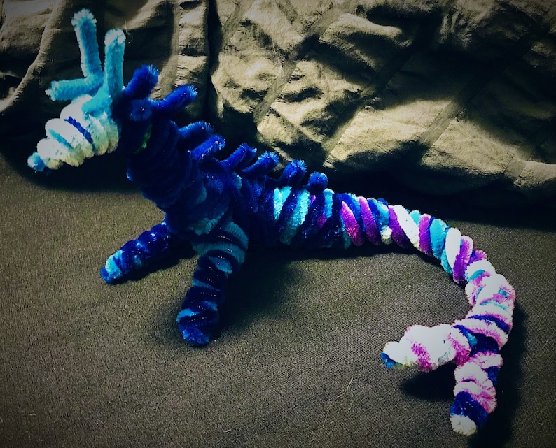 May include: A blue, purple, and white pipe cleaner dragon with a long tail. The dragon is curled up on a dark gray surface.