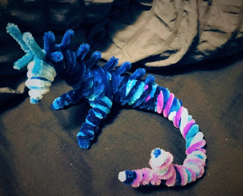 May include: A blue, pink, and white pipe cleaner dragon with a curled tail. The dragon is made of pipe cleaners and is a handmade craft.