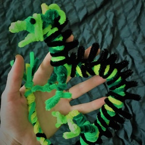 May include: A green and black pipe cleaner dragon with a long, curled tail. The dragon is made of pipe cleaners and has a green body with black stripes.