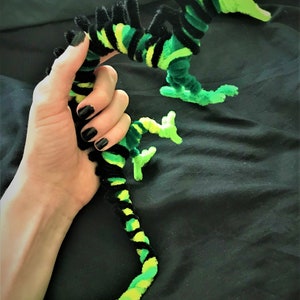 May include: A green and black pipe cleaner dragon with a long tail and a wide open mouth. The dragon is held in a person's hand.