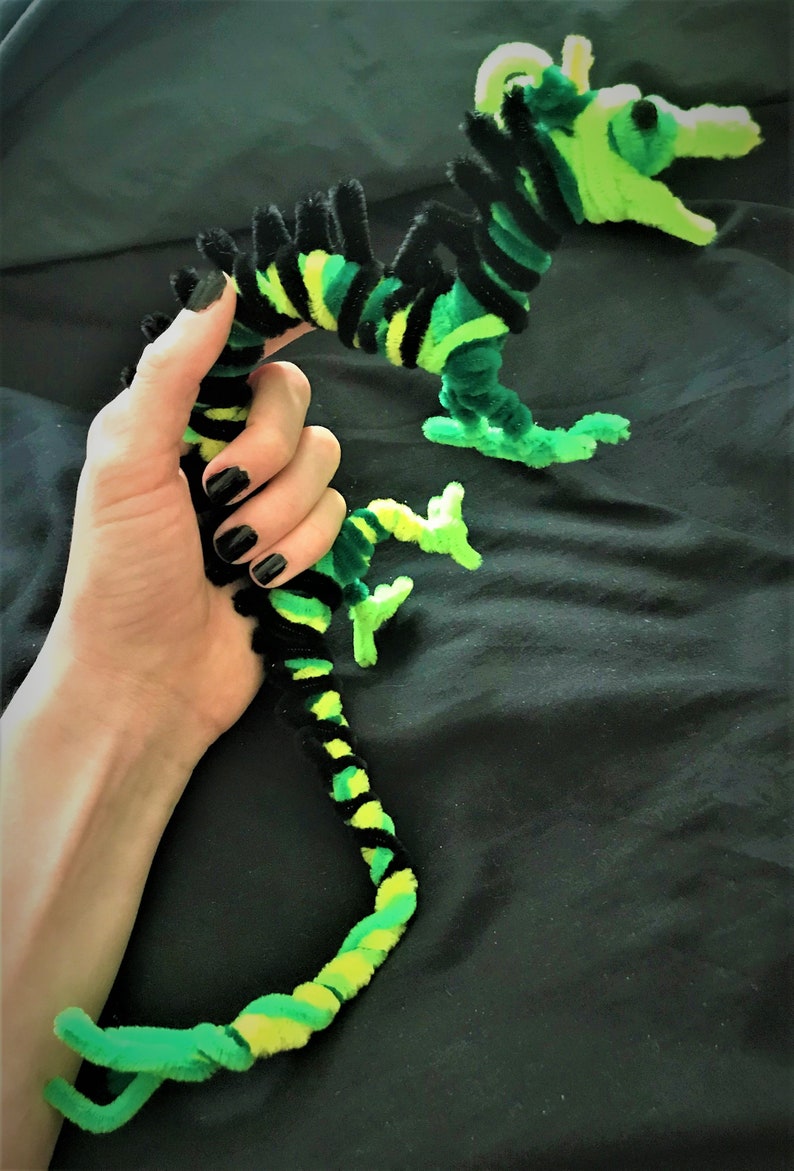 May include: A green and black pipe cleaner dragon with a long tail and a wide open mouth. The dragon is held in a person's hand.