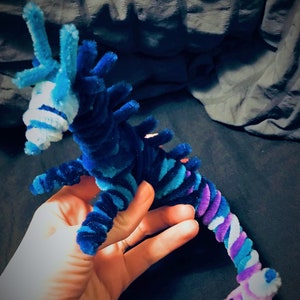 May include: A blue and purple pipe cleaner seahorse with white accents. The seahorse is made of pipe cleaners and is a handmade craft.