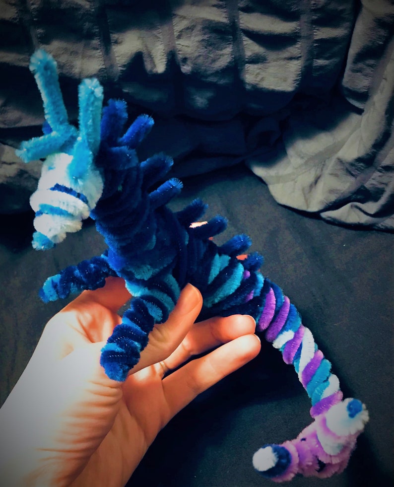 May include: A blue and purple pipe cleaner seahorse with white accents. The seahorse is made of pipe cleaners and is a handmade craft.