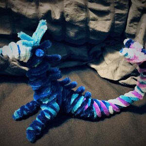 May include: A blue and purple pipe cleaner dragon with a white and blue head. The dragon is curled up in a sitting position with its tail curled around its body.