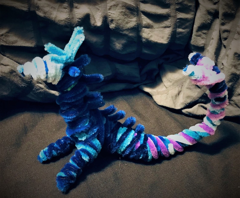 May include: A blue and purple pipe cleaner dragon with a white and blue head. The dragon is curled up in a sitting position with its tail curled around its body.