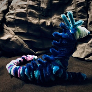 May include: A blue, green, and purple pipe cleaner creature with a long, segmented body and a head with three pipe cleaner antennae. The creature is lying on a dark surface.