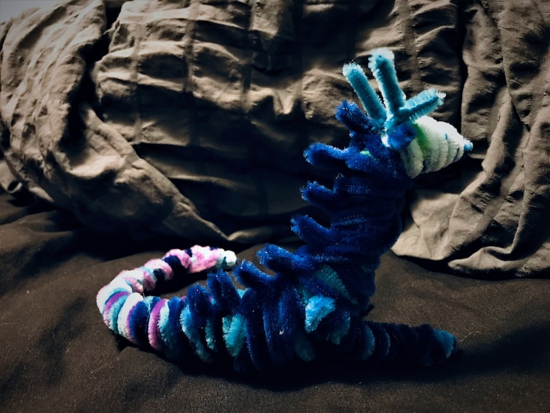 May include: A blue, green, and purple pipe cleaner creature with a long, segmented body and a head with three pipe cleaner antennae. The creature is lying on a dark surface.