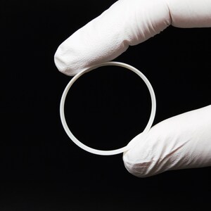 May include: A white, circular object is held by a gloved hand against a black background. The object appears to be a ring or a circular component, possibly made of plastic or a similar material. The image is well-lit, highlighting the object's shape.