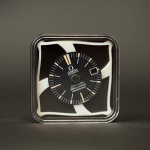 Puede incluir: Una esfera de reloj en blanco y negro con un marco de plástico transparente. La esfera del reloj tiene un fondo negro con marcas blancas y el texto "OMEGA AUTOMATIC Seamaster 600m/2000ft PROFESSIONAL".