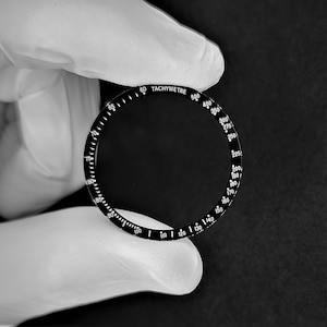 May include: Black bezel with white markings for a watch. The bezel has a tachymeter scale with numbers from 60 to 500.
