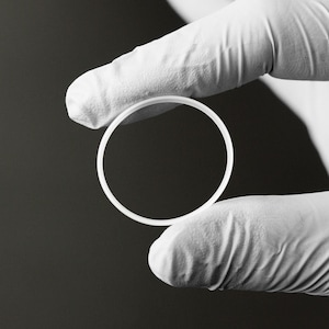 Può includere: Un oggetto circolare bianco è tenuto tra dita guantate su uno sfondo scuro. L'oggetto sembra essere un anello o una rondella, con una superficie liscia e pulita. L'immagine è ben illuminata, evidenziando la forma e la consistenza dell'oggetto.