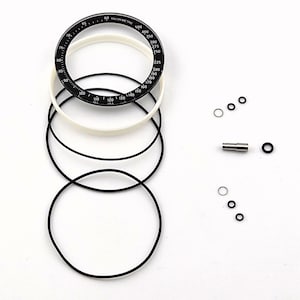 Puede incluir: Un inserto de bisel de taquímetro negro con marcas blancas, un anillo circular blanco, tres juntas circulares negras delgadas, un pequeño pasador cilíndrico de metal plateado y varios anillos circulares diminutos en negro y plata.