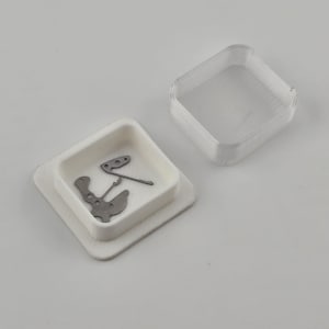 May include: A white square container with a lid, holding small metal watch parts. The parts are silver and have intricate shapes. The container is on a white surface.