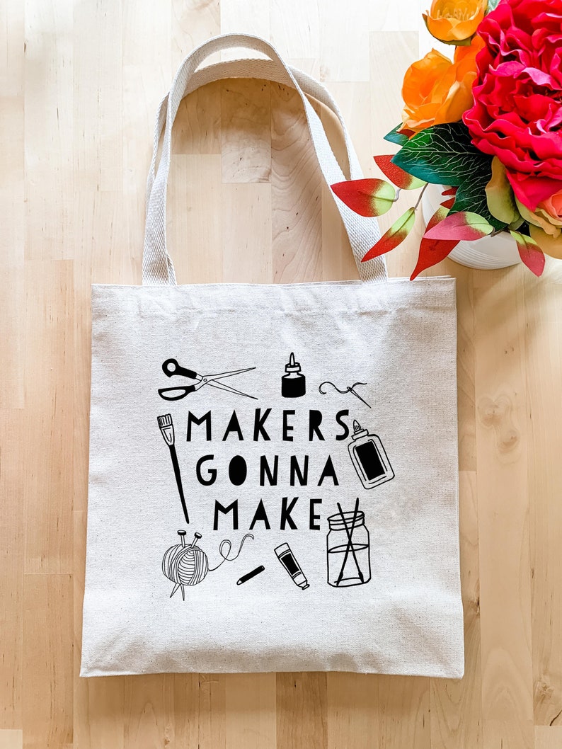 A Natural Canvas Bag Screen-printed Tote on a light wood table. Black images of scissors, paint brushes, glue, yarn and other crafting accessories surround the black text that reads "Makers Gonna Make."