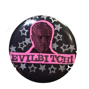May include: A round pinback button featuring a black background with silver glitter stars. In the center is a black and white portrait of a person outlined in pink, positioned above a pink horizontal banner with the black text EVILBITCH!