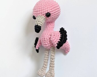 May include: A pink crocheted flamingo toy with black eyes and a black beak. The flamingo has black and white crocheted wings and long, thin legs.