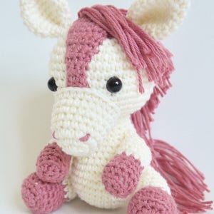 May include: A crocheted pink and white horse toy with black eyes. The horse is sitting with its legs tucked under its body. The horse has a pink mane and tail.