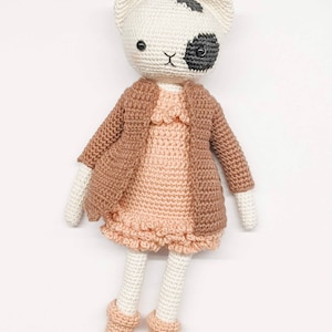 May include: A crocheted cat doll wearing a brown cardigan and a pink dress. The doll has black and white markings on its face and black eyes.