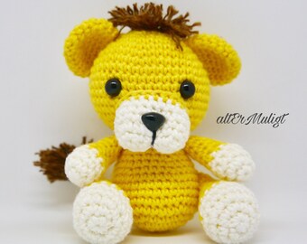 Puede incluir: Un león amarillo de crochet con una melena marrón, patas blancas y ojos negros. El león está sentado sobre una superficie blanca.