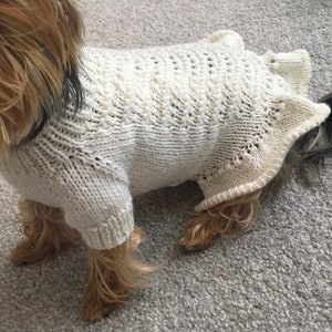 May include: A white knitted dog sweater with a delicate openwork pattern. The sweater is designed for small dogs and has a loose fit.