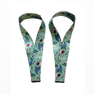 May include: Two floral patterned lanyards. The lanyards are a light green color with a repeating pattern of red, blue, and pink flowers and green leaves. The lanyards are made of a ribbon-like material and have a black border at the bottom.