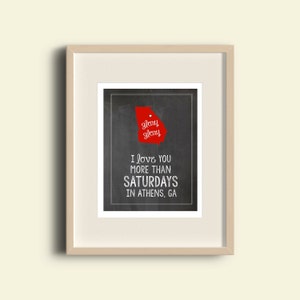 May include: Framed print with a red outline of Georgia, the text "glory glory", and the words "I love you more than Saturdays in Athens, GA" in white on a dark background. The frame is beige.