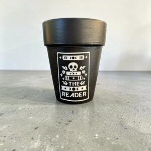 May include: A matte black tapered plant pot featuring a white graphic design of a skull resting on a stack of books. The design includes celestial moon phases, floral accents, and the text THE READER printed in a bold, clean, sans-serif font.