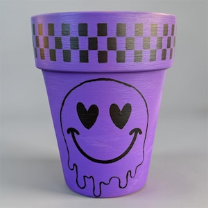 May include: A purple plant pot featuring a black checkerboard pattern around the top rim. The center of the pot displays a black graphic of a melting smiley face with heart-shaped eyes, painted in a bold, clean style on the smooth purple surface.