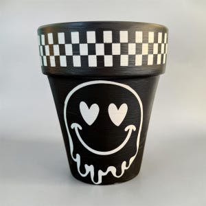 May include: A black terracotta plant pot featuring a white checkerboard pattern around the upper rim. The front of the pot displays a large white graphic of a melting smiley face with heart-shaped eyes, painted in a bold, high-contrast style.
